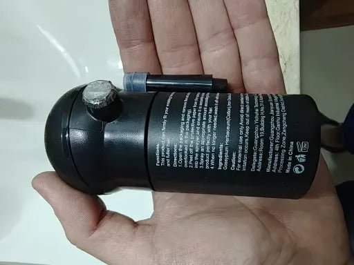 Um frasco preto do produto é exibido horizontalmente sobre a palma da mão.